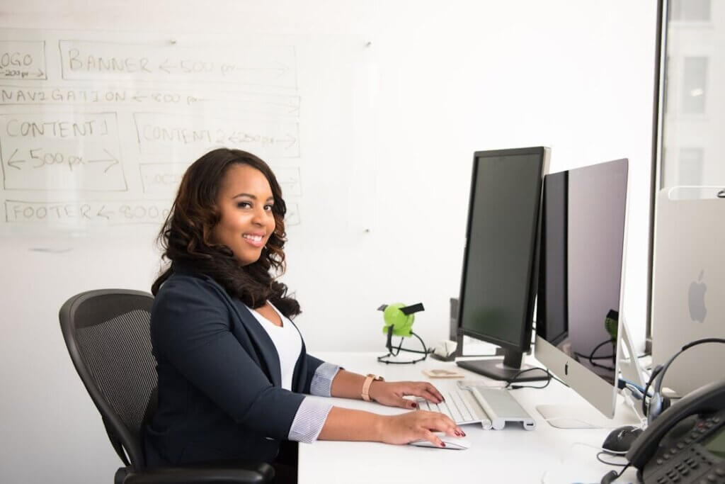 Le leadership féminin en entreprise Femme dirigeante concentrée travaillant sur ordinateur au bureau moderne, avec tableau stratégique et double écran, illustrant le leadership féminin en action et la gestion professionnelle efficace.